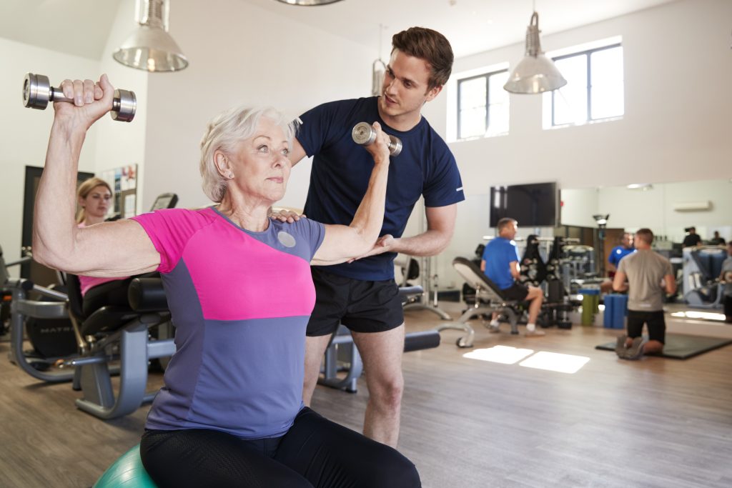 A personal trainer supports a woman in the gym to lift weights as part of her pain management programme.