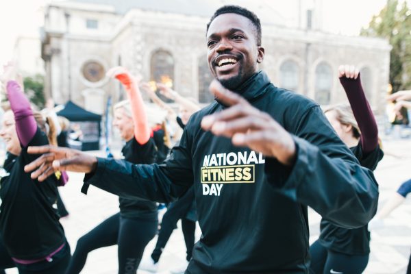 Group of women and one man exercising in a town square, wearing t-shirts saying National Fitness Day