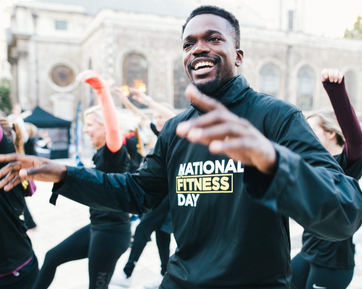 Group of women and one man exercising in a town square, wearing t-shirts saying National Fitness Day