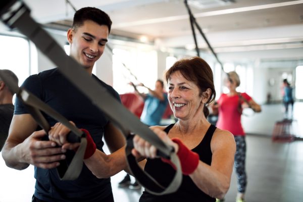 A group of cheerful seniors in gym with a young trainer doing exercise with TRX.