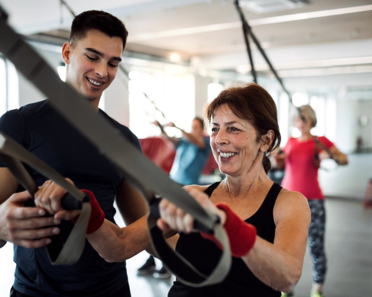 A group of cheerful seniors in gym with a young trainer doing exercise with TRX.
