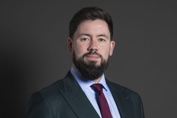 Portrait of Adam Thompson MP, wearing a black suit, blue shirt and purple tie.