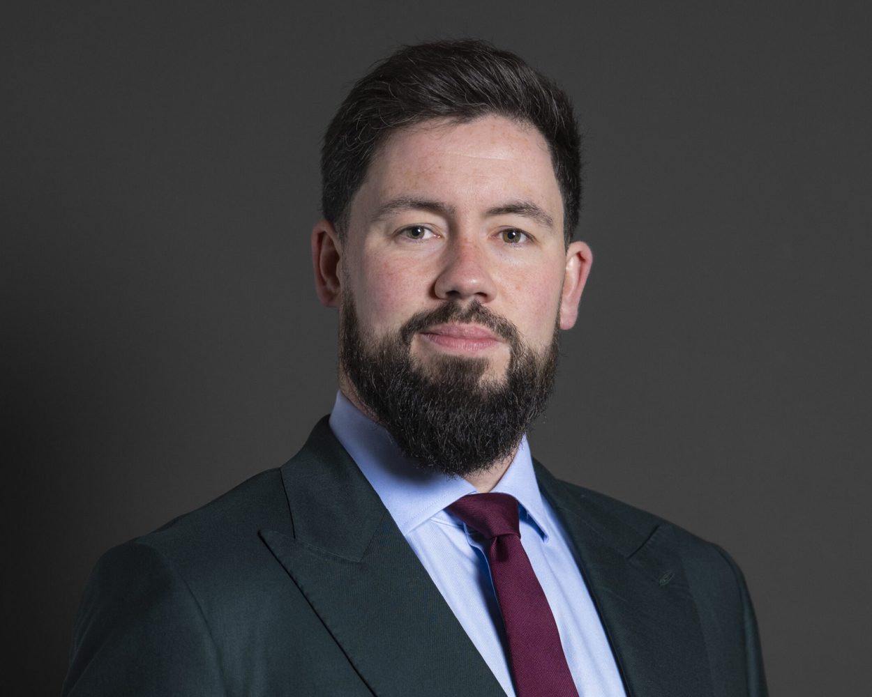 Portrait of Adam Thompson MP, wearing a black suit, blue shirt and purple tie.