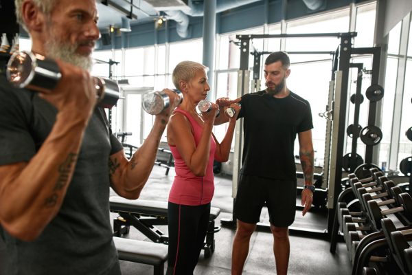 Concentrated and determined senior woman and man with dumbbells excercising together with personal trainer. Coach adjusting workout according to client's physical condition.