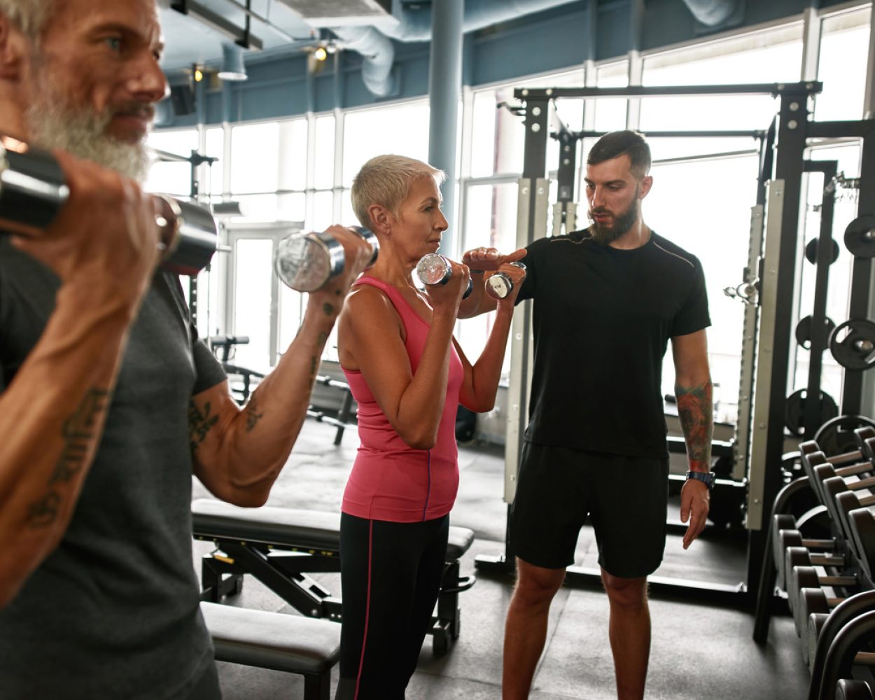 Concentrated and determined senior woman and man with dumbbells excercising together with personal trainer. Coach adjusting workout according to client's physical condition.
