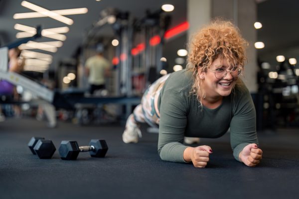 Determined smiling young woman gym fitness client with curly hair in plank position doing exercises for strong core abdominal muscles having dumbbell weight set next to her focused on fitness goal at gym.