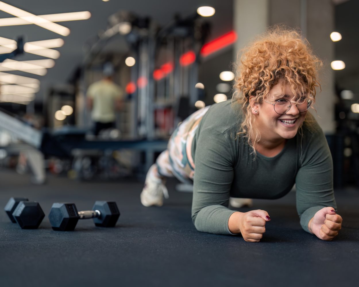 Determined smiling young woman gym fitness client with curly hair in plank position doing exercises for strong core abdominal muscles having dumbbell weight set next to her focused on fitness goal at gym.