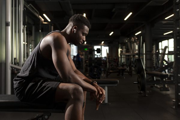 young athletic african american man sits in dark gym and thinks, pensive athletic guy rests and looks forward