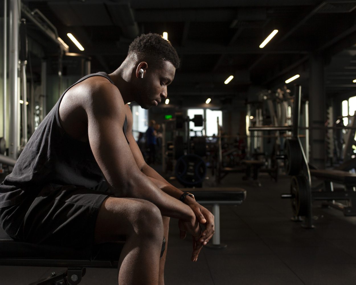 young athletic african american man sits in dark gym and thinks, pensive athletic guy rests and looks forward