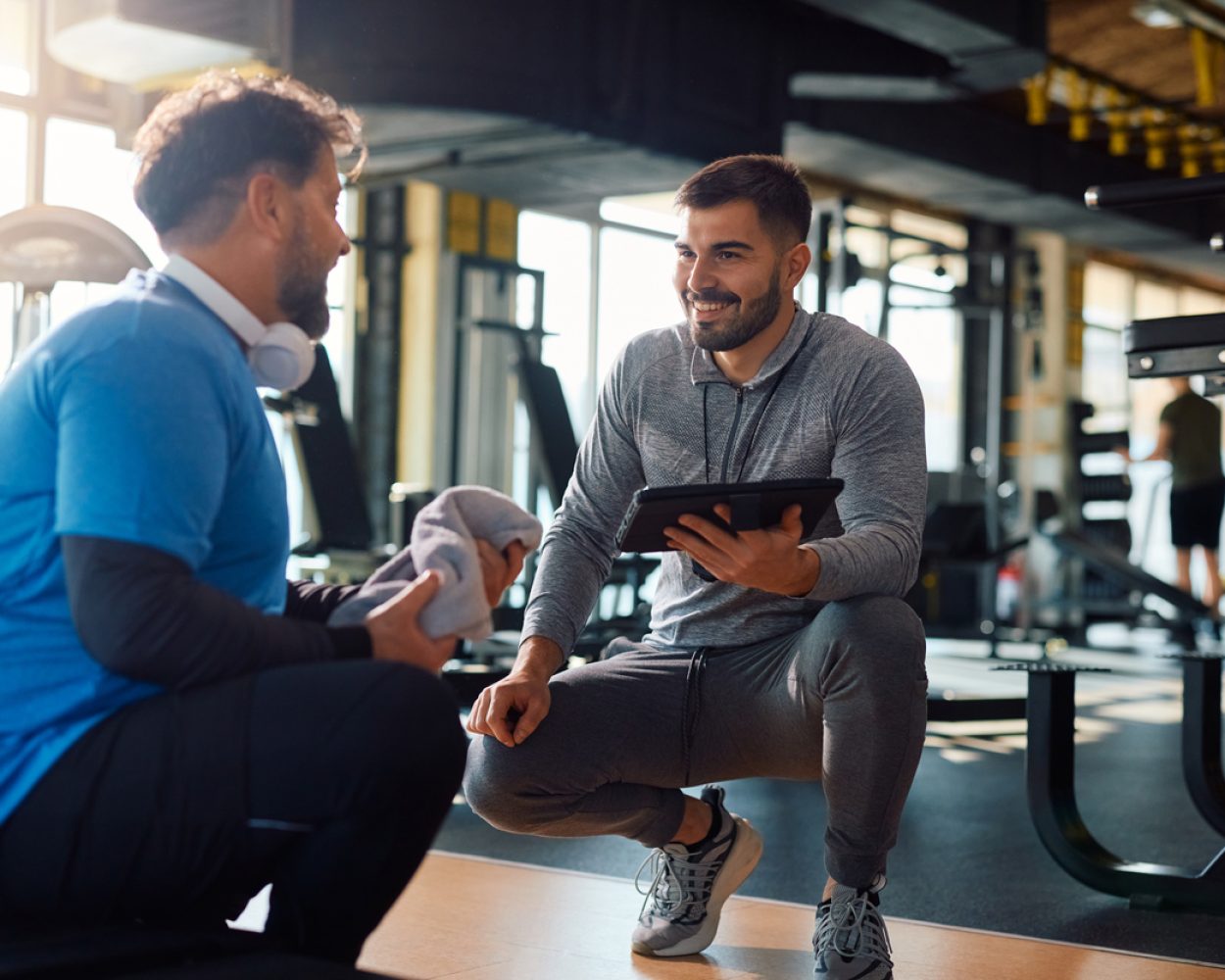 Fitness instructor using touchpad while talking to mature man during exercise class in a gym.