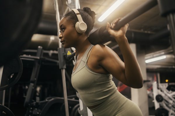 Strong black sportswoman with headphones squatting doing workouts on smith machine at gym.