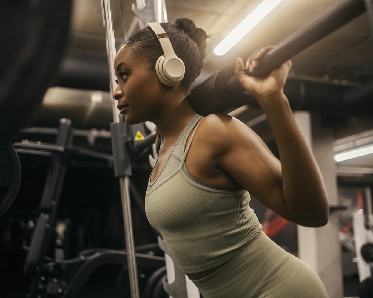 Strong black sportswoman with headphones squatting doing workouts on smith machine at gym.
