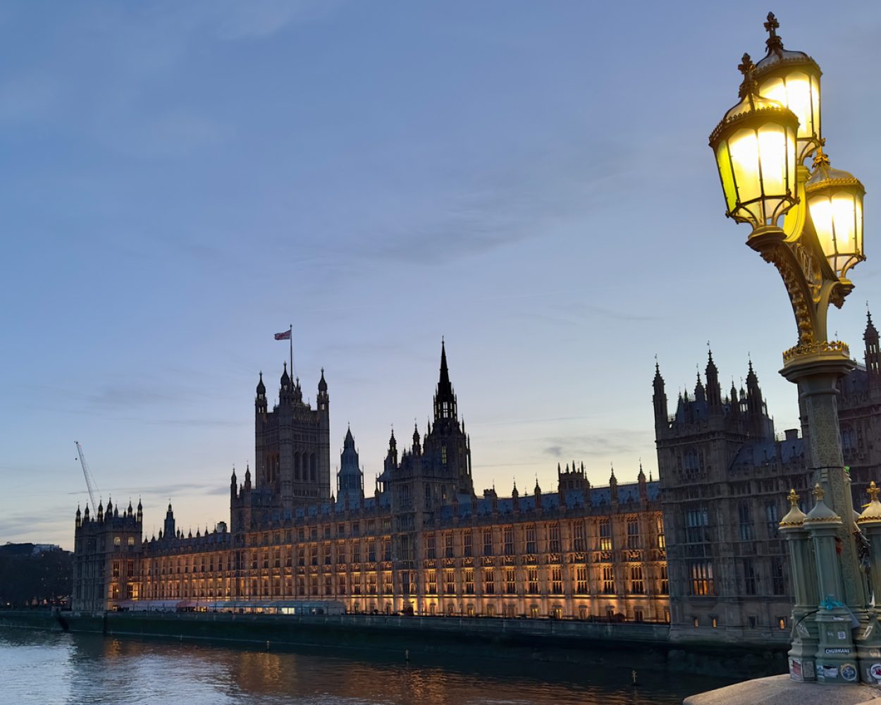The Houses of Parliament in Westminster, London at dusk with an historic lamp on Westminster Bridge in the foreground.