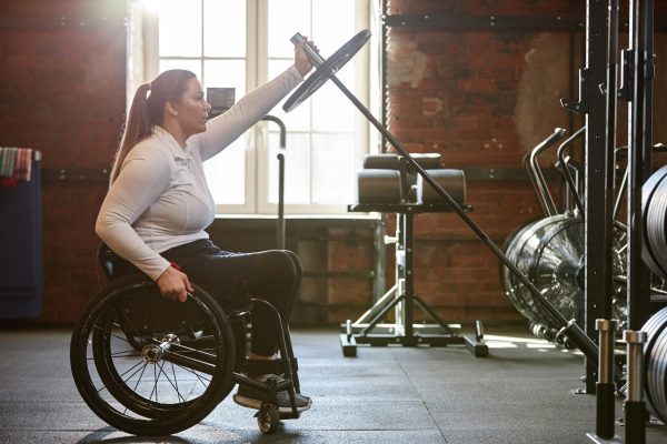 Side view portrait of adult woman with disability using wheelchair in gym and doing exercises with barbell enjoying arm workout copy space